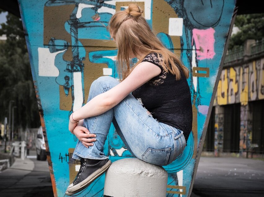 Ein jugendliches Mädchen hockt auf einem Poller mit abgewendetem Gesicht, im Hintergrund eine Graffiti-Wand