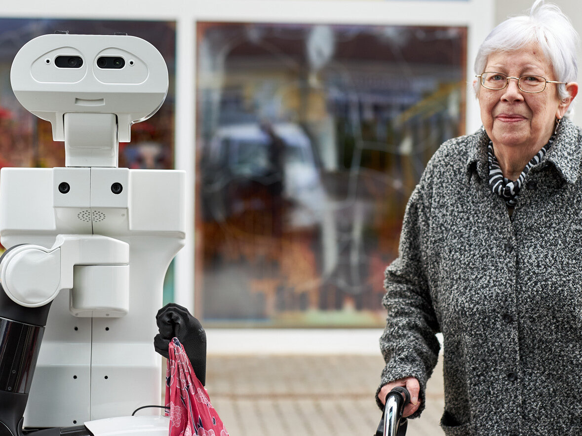 Eine ältere Dame und ein ihr assistierender Roboter stehen an der Straße