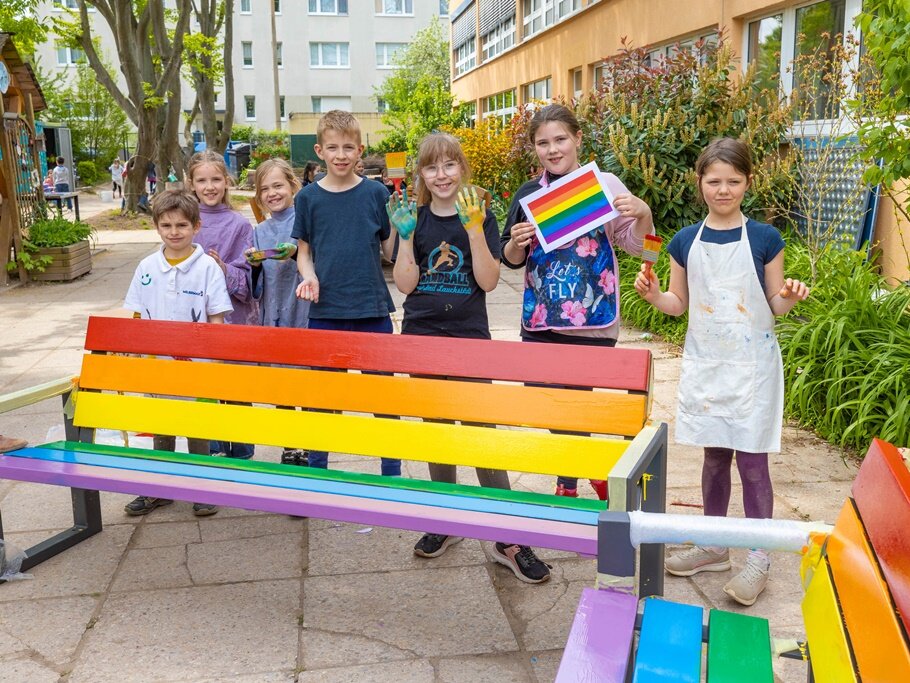 Hinter den regenbogenfarbigen Bänken stehen Kinder.